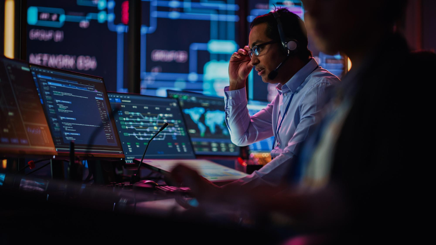 Two men working in a dark room with computer screens and headsets, immersed in their tasks